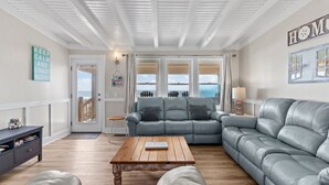 A living room with a grey leather sofa, a wooden coffee table, and a view of the ocean through large windows. - Oceanfront family fun home and dog friendly! Coastal Coop (Oak Island)