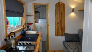 A compact kitchen with a gas stove, sink, and wooden accents. A small dining area with a patterned sofa is visible. - Unique Upcycled Straw Trailer @ Westcote (Hawick)