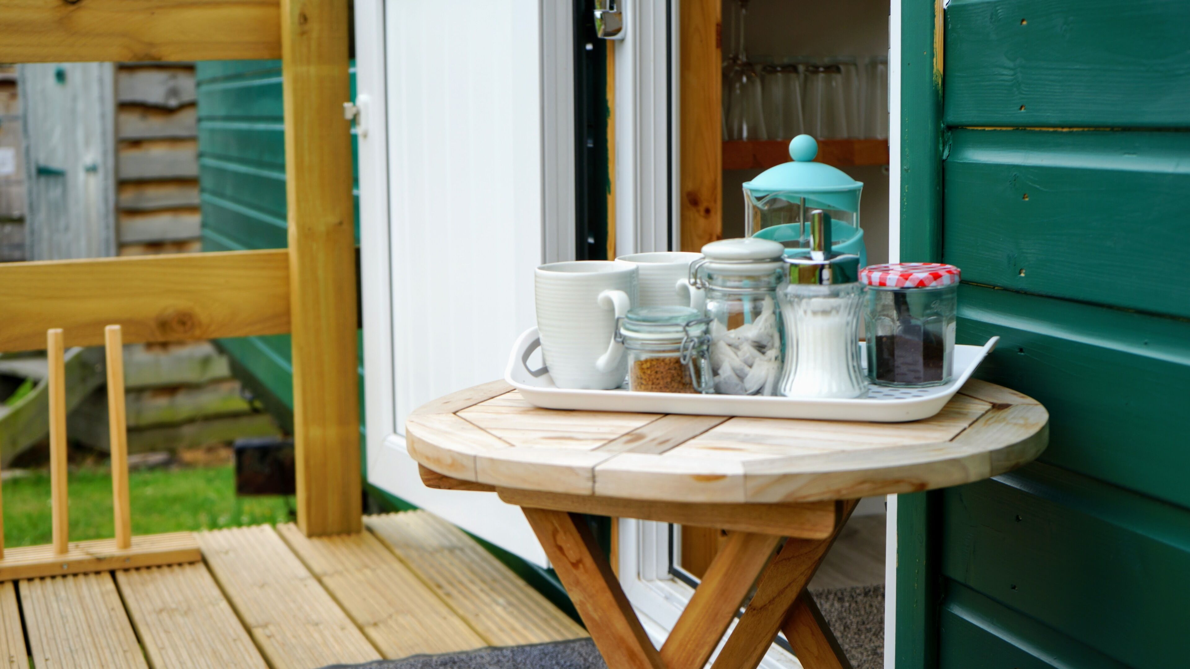A wooden table with a tray holding white mugs, a teapot, and jars on a porch with a wooden fence and a green wall in the background.