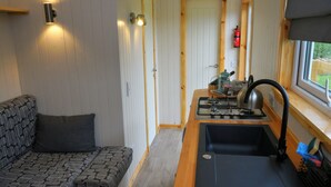 A compact kitchen with a stove, sink, and window. A sofa with a patterned cushion is visible in the foreground. - Unique Upcycled Straw Trailer @ Westcote (Hawick)