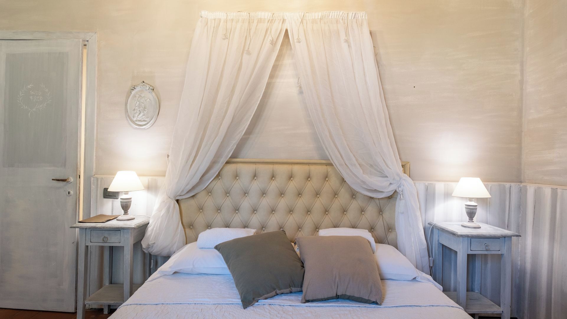 A bedroom with a tufted headboard, two bedside tables with lamps, a bed with white and brown pillows, and a door with a chalkboard sign.