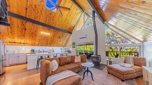 A spacious living area with a wooden ceiling, a fireplace, and a large window overlooking greenery. - Oakhurst Cabin|Bass Lake View|Game Room|Hot Tub! (Oakhurst)
