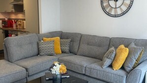 A grey sectional sofa with patterned cushions, a black coffee table with a vase of flowers, and a wall clock. - WENDY APARTMENT HOME AWAY FROM HOME (Aberdeen)