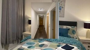 A hotel room with a bed, bedside tables, a nightstand, a painting on the wall, and a hallway leading to another room. - WENDY APARTMENT HOME AWAY FROM HOME (Aberdeen)