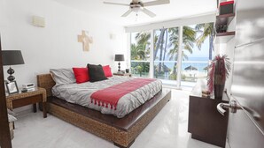 A modern bedroom with a large bed, a ceiling fan, a window with a view of palm trees and the ocean, and a small desk with a lamp. - Casa Caracol (Bucerías)