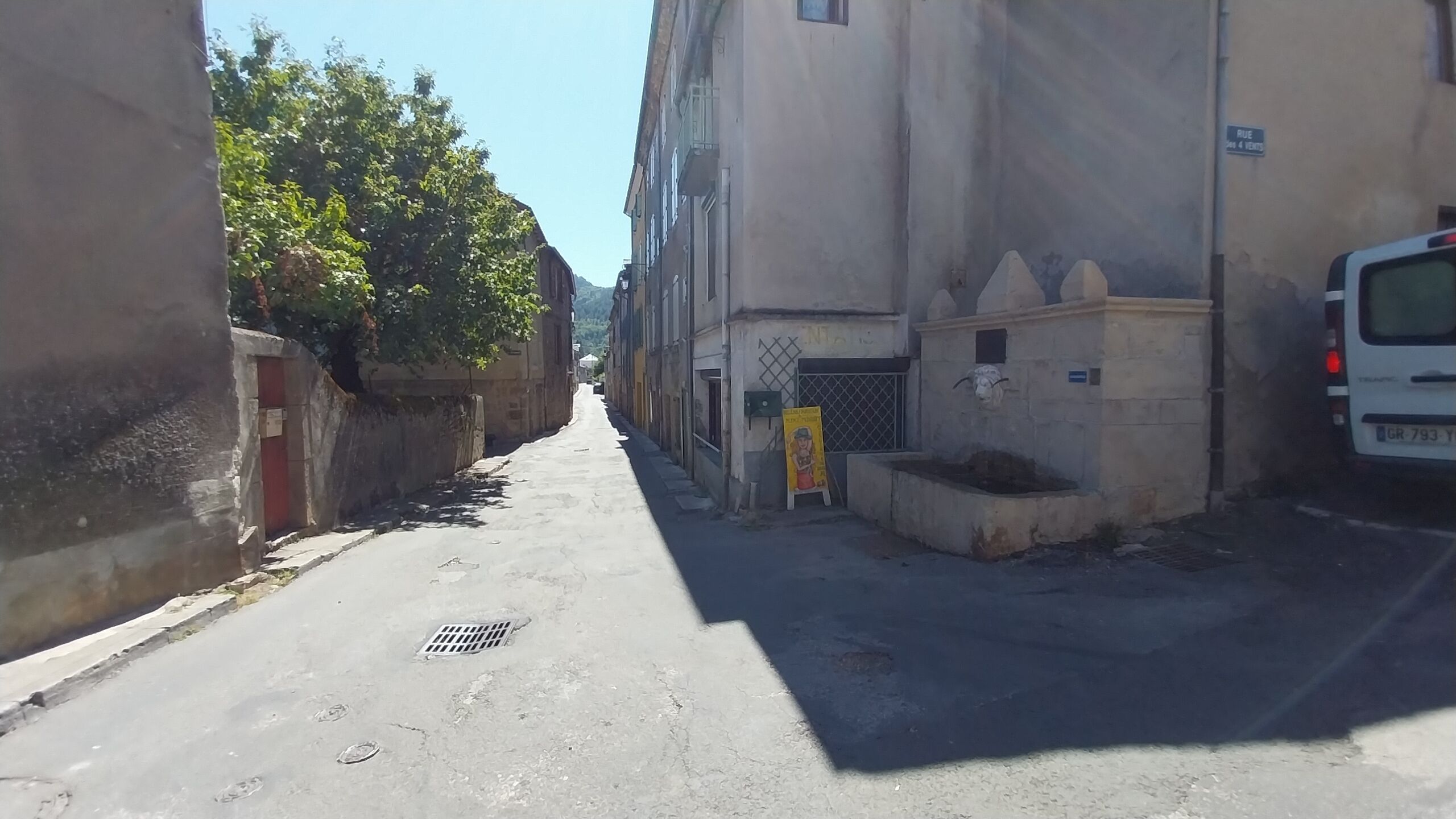 A narrow street with buildings on both sides, a white van parked on the right, and a signboard on the left.