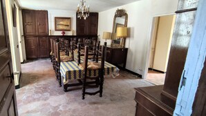 Un coin repas avec un sol carrelé en forme d’hexagone, des chaises en bois recouvertes de nappes à motifs, un lustre et un miroir au-dessus d’une commode.