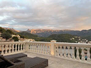 Balcó amb mobiliari de vimet i barana blanca que dona a un paisatge de muntanya amb edificis.