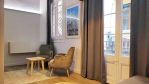 Habitació amb un llit, dues cadires, una taula petita, una finestra amb cortines i una prestatgeria penjada a la paret.