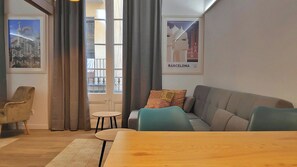 Ett modernt vardagsrum med en soffa, ett litet runt bord och ett matbord. Det finns gardiner, ett fönster med utsikt och två inramade tavlor på väggen.