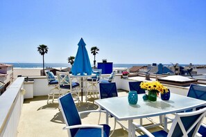 Terraza en la azotea con mesas blancas y sillas azules, con vista a una playa y el océano.