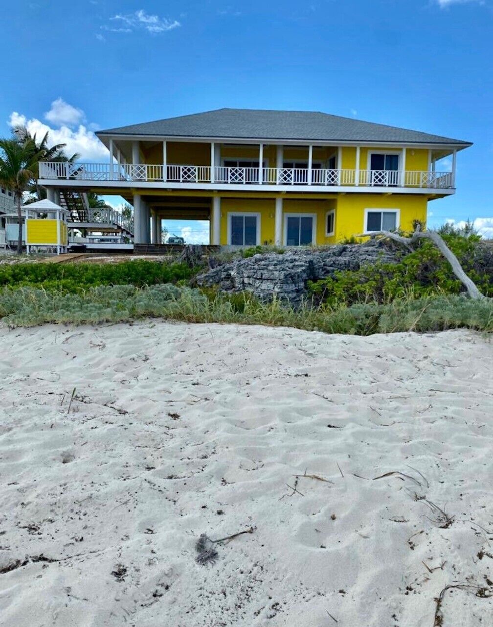 Una casa amarilla de dos pisos frente a la playa, con balcón, ubicada en una playa arenosa con vegetación al frente.