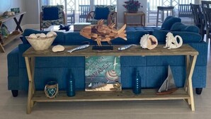 A spacious living room with a large blue sofa, a wooden coffee table, and a ceiling fan. There are two large windows with a view of the ocean. - Beautiful Casa Sol  (Great Harbour Cay)