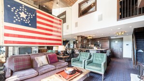 A spacious living room with a large American flag, a cozy fireplace, and a modern kitchen in the background. - Western Cowboy Quarters steps away from Vail Gondola w/ Hot tub  and  Spa access (Vail)