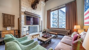 A cozy living room with a stone wall, a mounted deer head, a flat-screen TV, a fireplace, and a large window with a view of a building. - Western Cowboy Quarters steps away from Vail Gondola w/ Hot tub  and  Spa access (Vail)