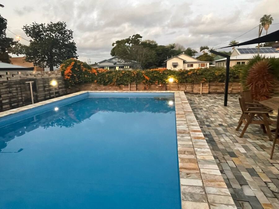 A rectangular swimming pool with a stone border, surrounded by a patio area with a table and chairs.