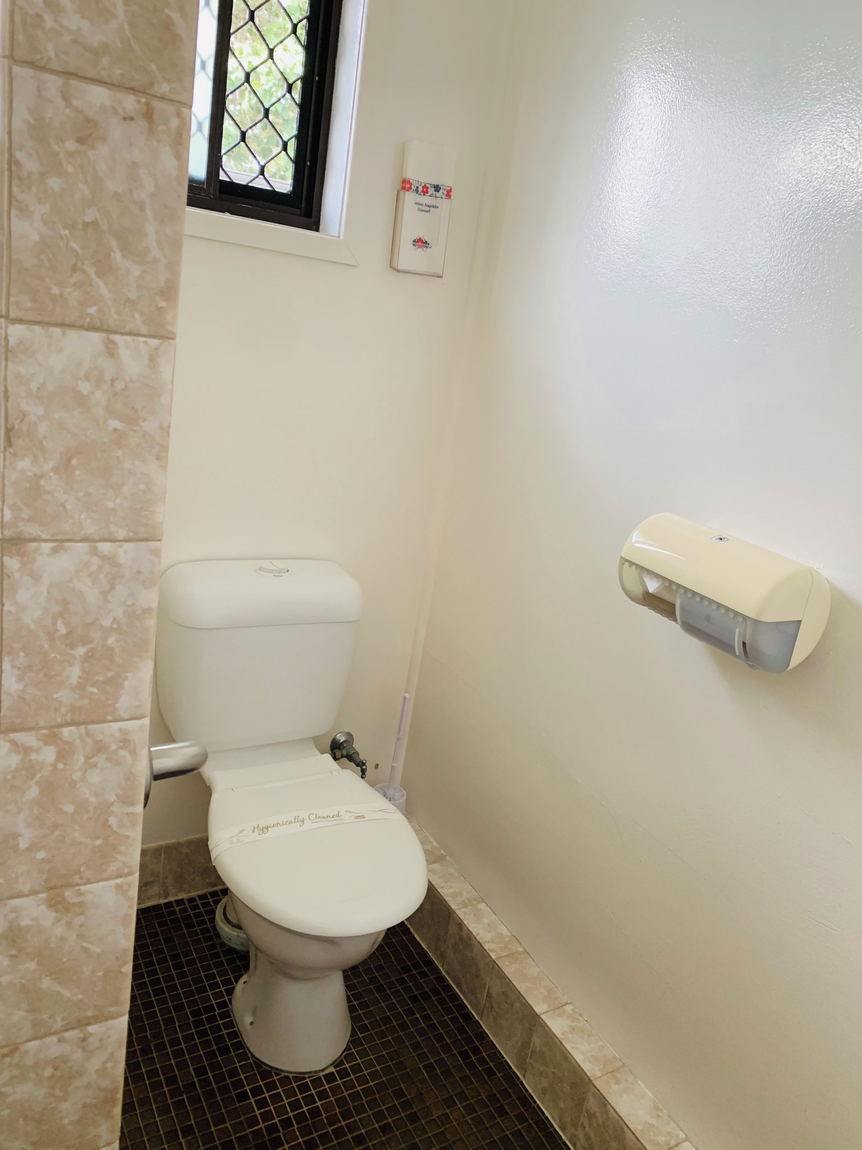 Une petite salle de bain propre, équipée de toilettes, d’un distributeur de gel hydroalcoolique et d’une fenêtre avec un moustiquaire en métal.