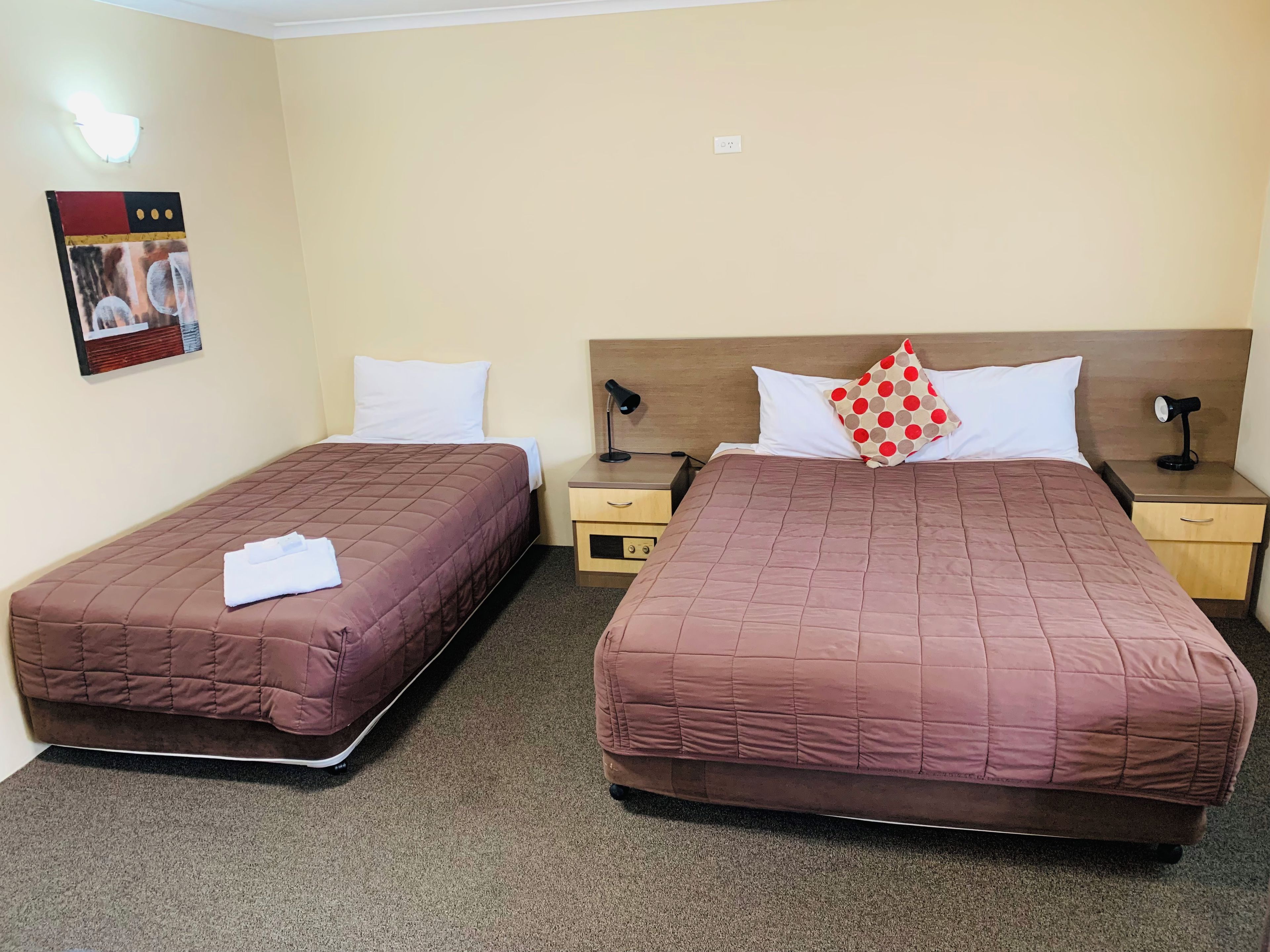 Ein Hotelzimmer mit zwei Betten, jeweils mit einer braunen gesteppten Tagesdecke, weißen Kissen und einem roten gepunkteten Kissen auf einem Bett. Es gibt Nachttische mit Lampen, eine Wandleuchte und einen kleinen Bilderrahmen.