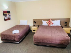 Habitación de hotel con dos camas, cada una con cubrecama acolchada marrón, almohadas blancas y una almohada roja con lunares en una cama. Hay mesitas de noche con lámparas, un aplique de pared y un pequeño marco de fotos.