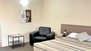 A hotel room with a bed, a chair, a small table, and a wall-mounted lamp. - Milgate Motel (Casino)