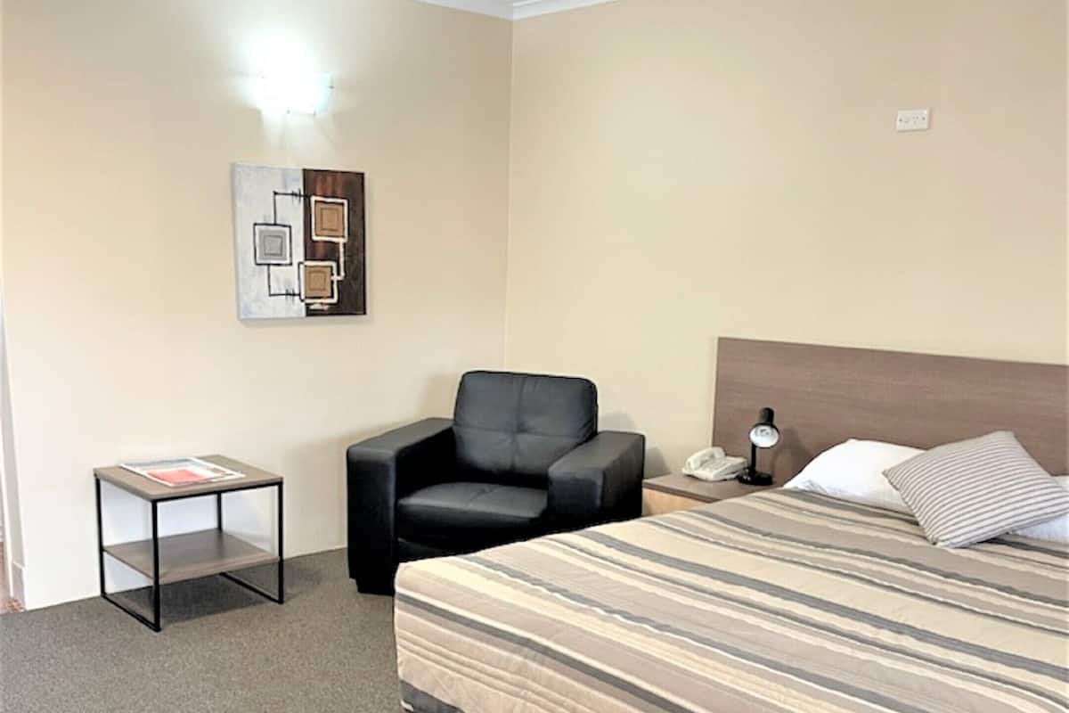 A hotel room with a bed, a chair, a small table, and a wall-mounted lamp.