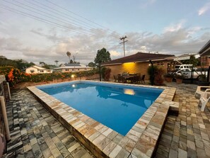 A rectangular swimming pool with a tiled edge, surrounded by a patio area with stone tiles.