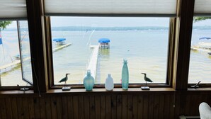A room with a large window overlooking a dock and water. There are two birds on the windowsill, and several bottles and a small figurine are placed on the windowsill.