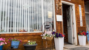 Un bâtiment en bois avec une pancarte « Bienvenue », une cloche et une pancarte « Bureau ». On peut voir des pots de fleurs et une jardinière suspendue à l’extérieur.