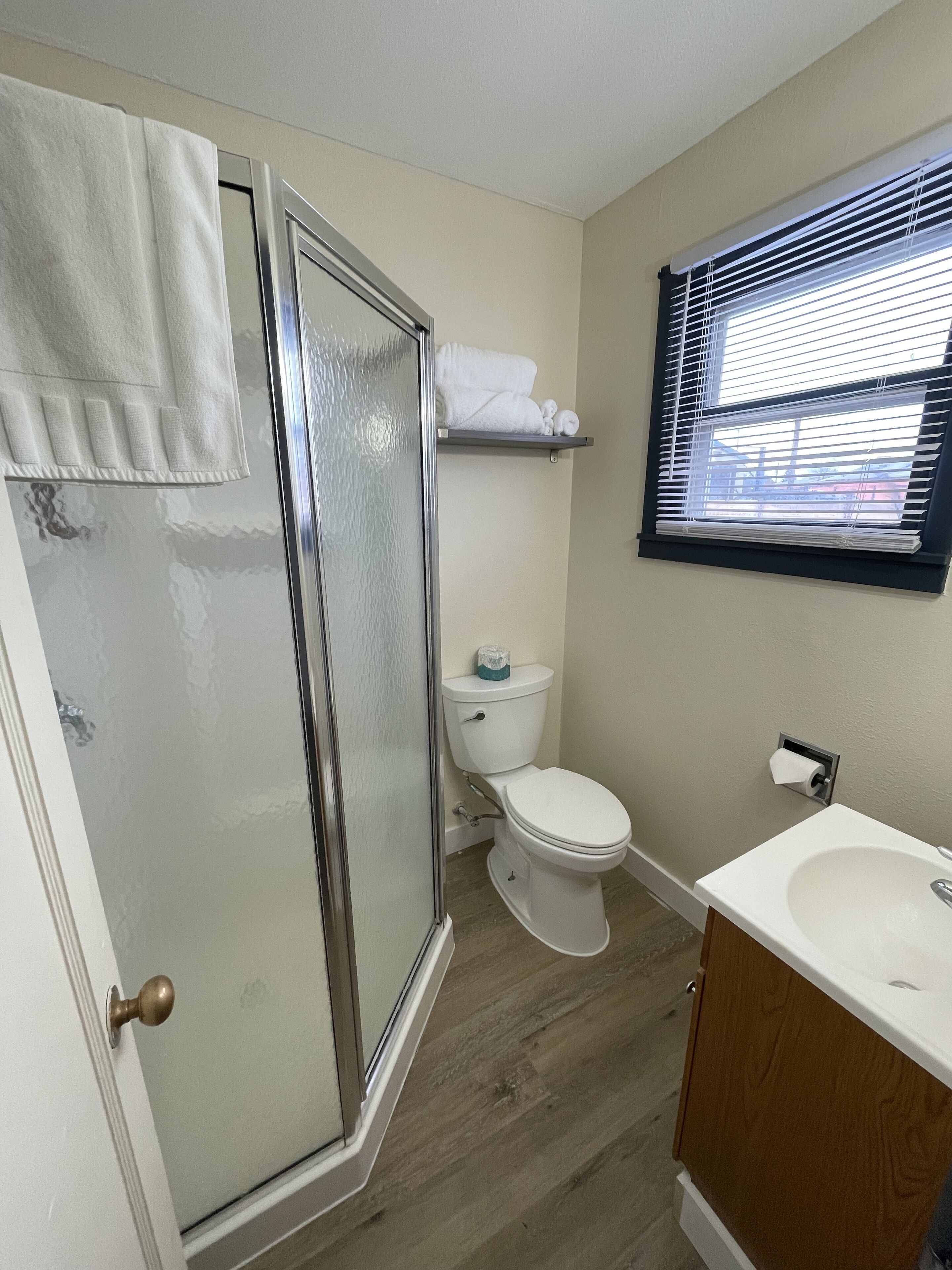 Une salle de bain avec une porte de douche en verre, des toilettes, un lavabo et une fenêtre avec des stores.