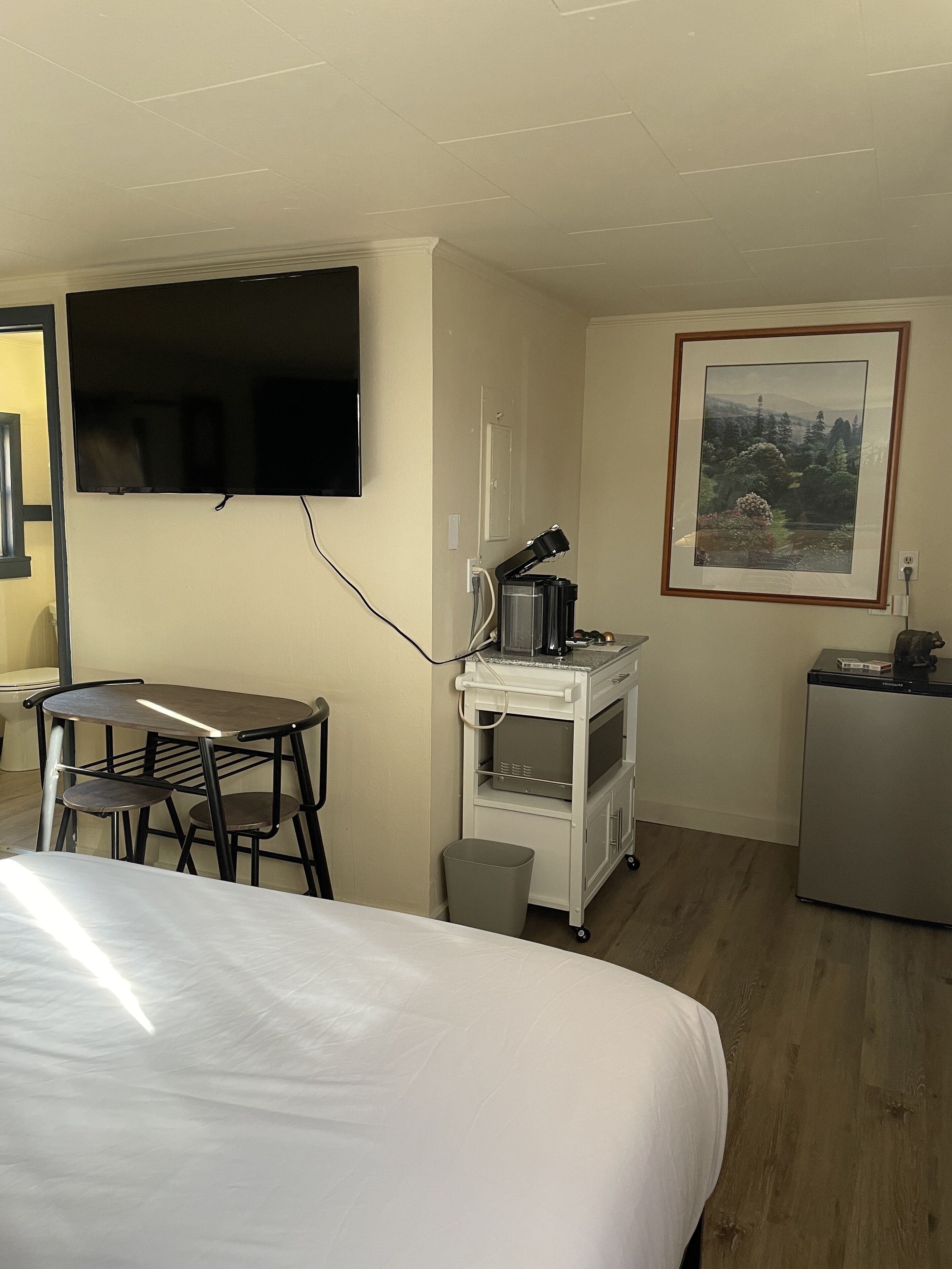 A hotel room with a bed, a desk with chairs, a TV, a kitchenette, and a framed picture on the wall.