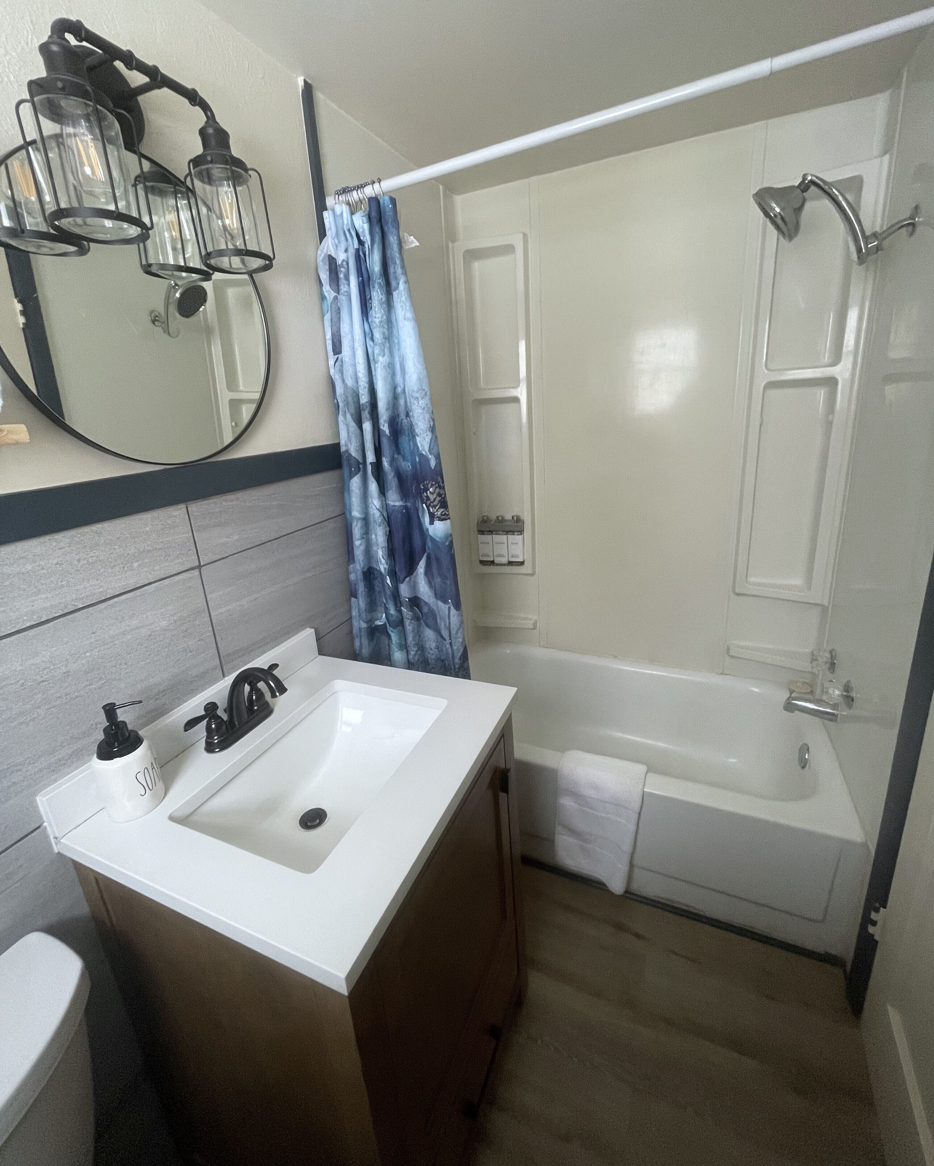 Un bagno con un lavandino bianco, un rubinetto nero e uno specchio sopra. Tende da doccia con motivo blu, una vasca da bagno bianca e un asciugamano appeso al muro.