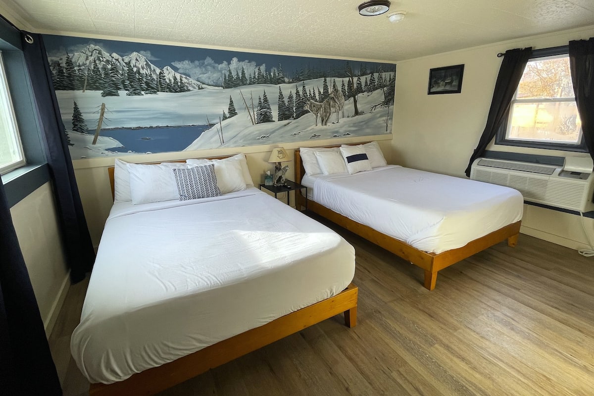 A room with two beds, a mural of a snowy landscape, a window with curtains, and a radiator.