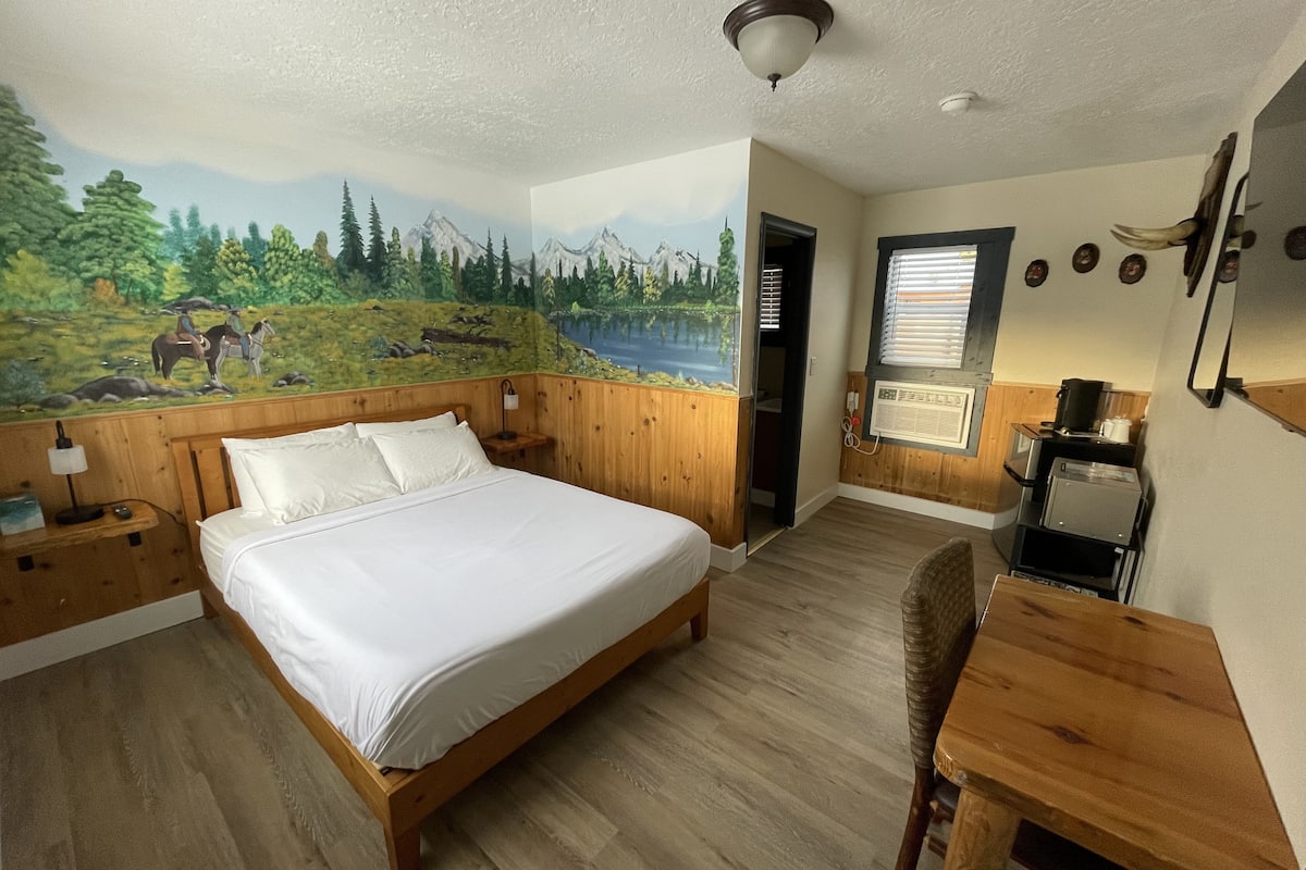A room with a bed, wooden headboard, a mural of a landscape, a small table, and a window with blinds.