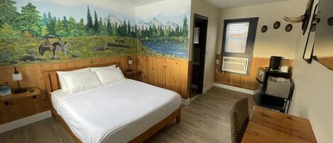 A room with a bed, wooden headboard, a mural of a landscape, a small table, and a window with blinds.