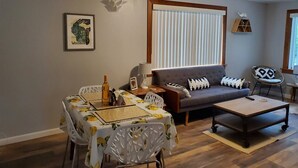 A living room with a sofa, a dining table with chairs, a coffee table, and a window with blinds. - The Cherry Pit - New listing (Sturgeon Bay)