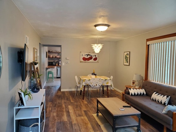 Un salon comprenant un canapé, une table basse et un coin repas avec une table et des chaises. Un téléviseur est fixé au mur et un luminaire est suspendu au plafond.