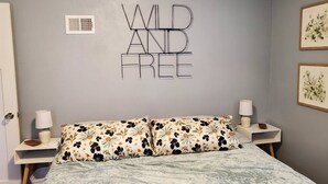 A bedroom with a bed, floral-patterned pillows, a wall-mounted sign saying "WILD AND FREE", a white bedside table with a lamp, and a framed picture on the wall. - The Cherry Pit - New listing (Sturgeon Bay)