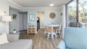 Una sala de estar moderna con un sofá gris, un sillón azul, una zona de comedor con una mesa y sillas blancas, y un gran ventanal con vistas a árboles.