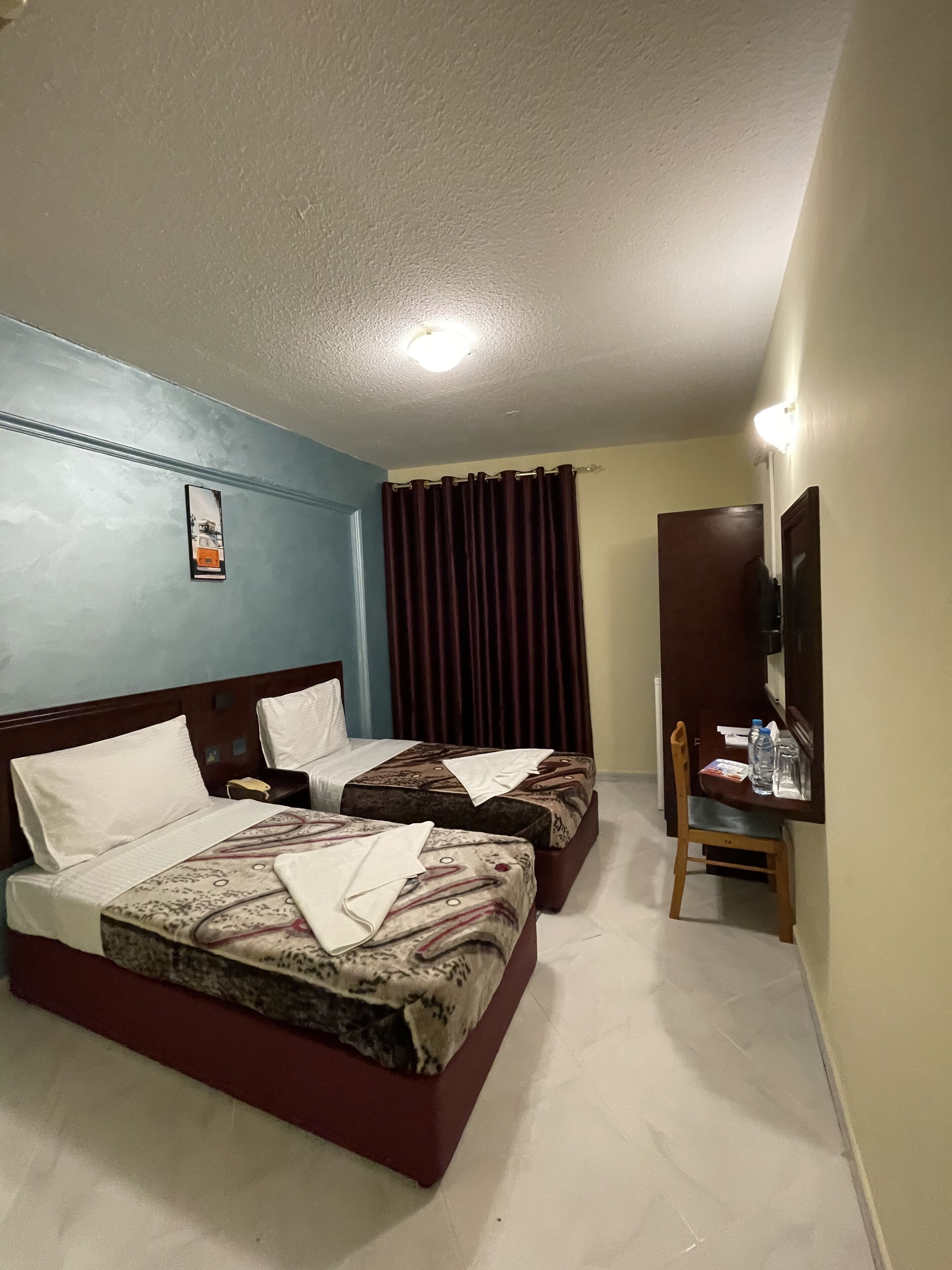 A hotel room with two beds, a wooden desk, a chair, a television, and a curtain.