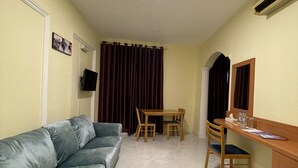 A compact living space with a grey sofa, a wooden dining table with chairs, a small desk with a chair, and a TV mounted on the wall. - MOUNT SINA HOTEL BY AURA (Dubai)