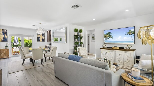 Ein modernes Wohnzimmer mit grauem Sofa, Essbereich und Fernseher, der eine Strandansicht zeigt.