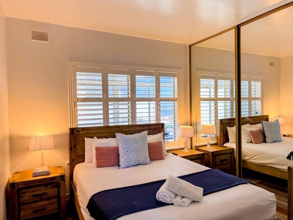 A hotel room with a large bed, wooden headboard, two bedside tables, a mirror, and a window with blinds.