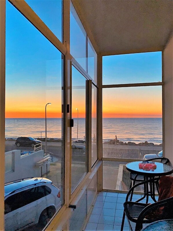A balcony with a view of the ocean at sunset, a table with chairs, and parked cars.