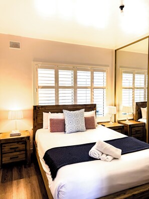 A hotel room with a large bed, wooden headboard, two bedside tables with lamps, a window with blinds, and a mirror on the wall.