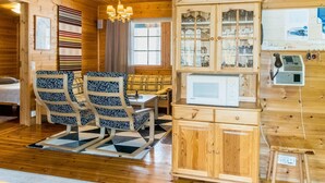 Interior acogedor de una cabaña con paredes de madera, un comedor con sillas estampadas, un televisor empotrado y un mueble de madera con puertas de cristal.