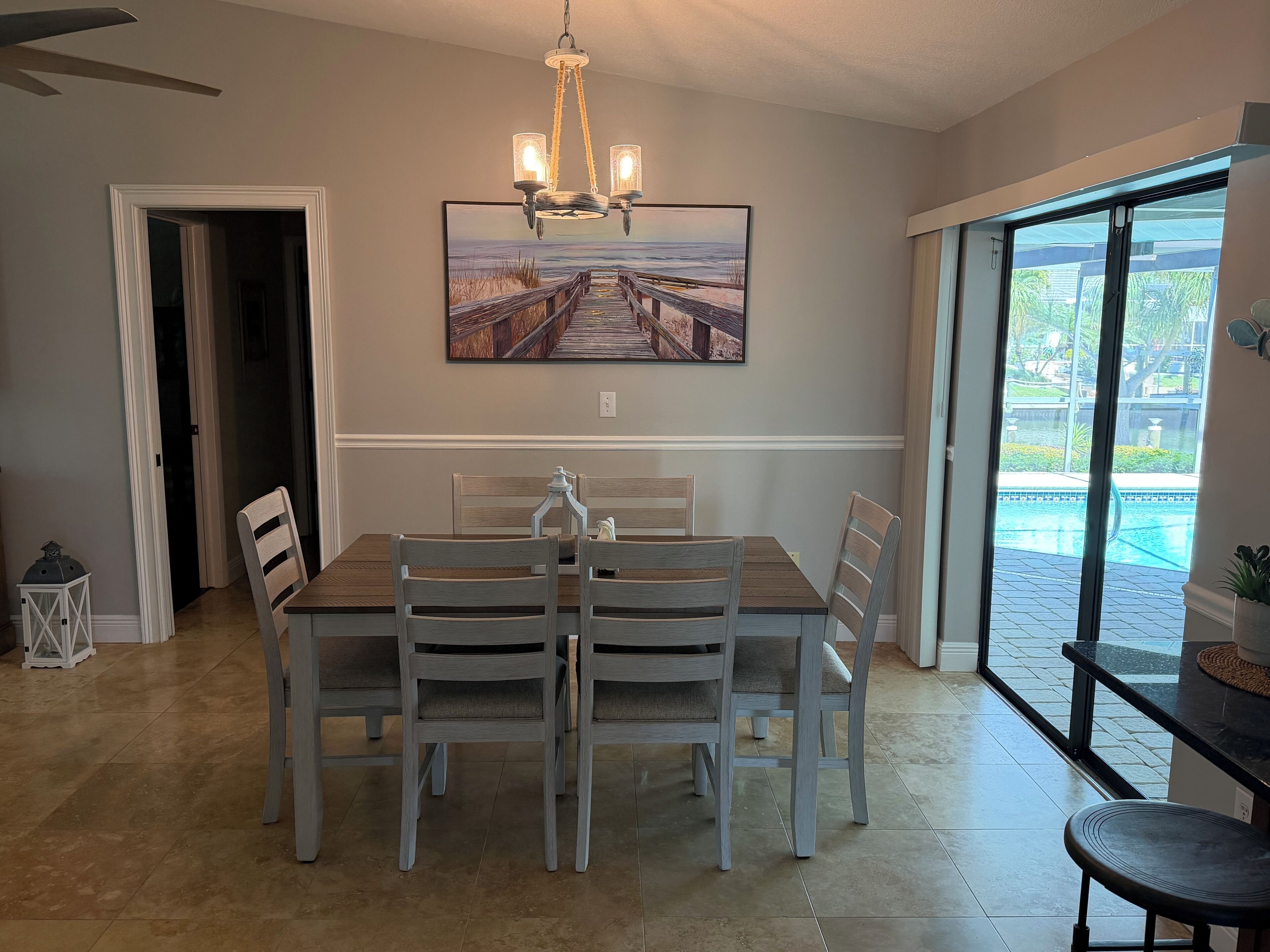 Un espace repas avec une table et des chaises en bois, un lustre, un grand tableau représentant une jetée et une vue sur une piscine, visible à travers de grandes fenêtres.