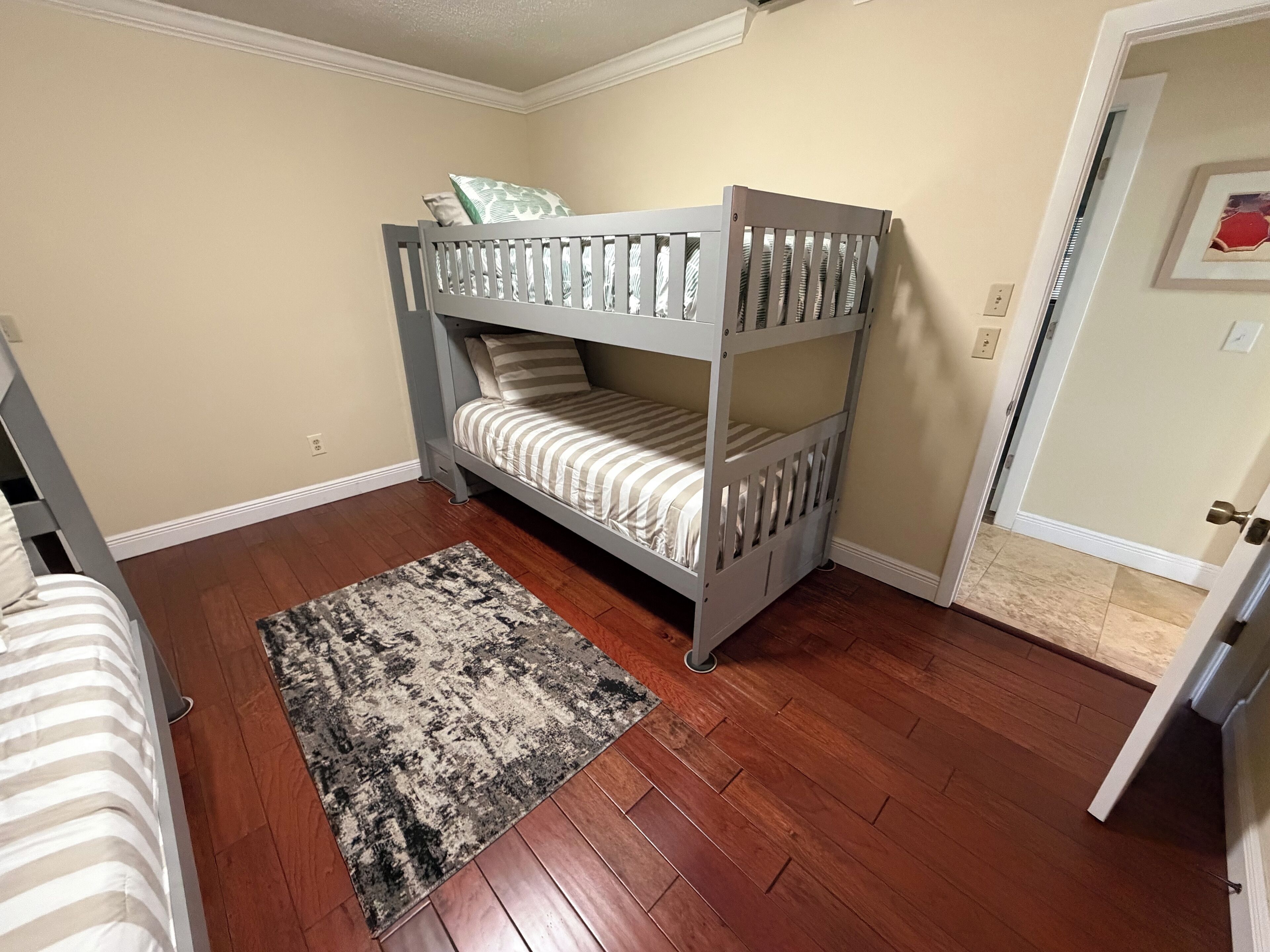 Una cama litera con ropa de cama a rayas, un tapete estampado y suelo de madera en una habitación con paredes color beige.
