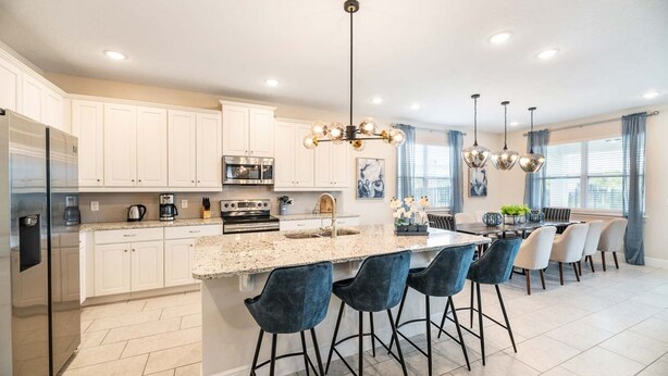 A modern kitchen with white cabinets, a stainless steel refrigerator, a built-in oven, and a granite countertop. There are four bar stools with dark upholstery and a dining table with chairs.
