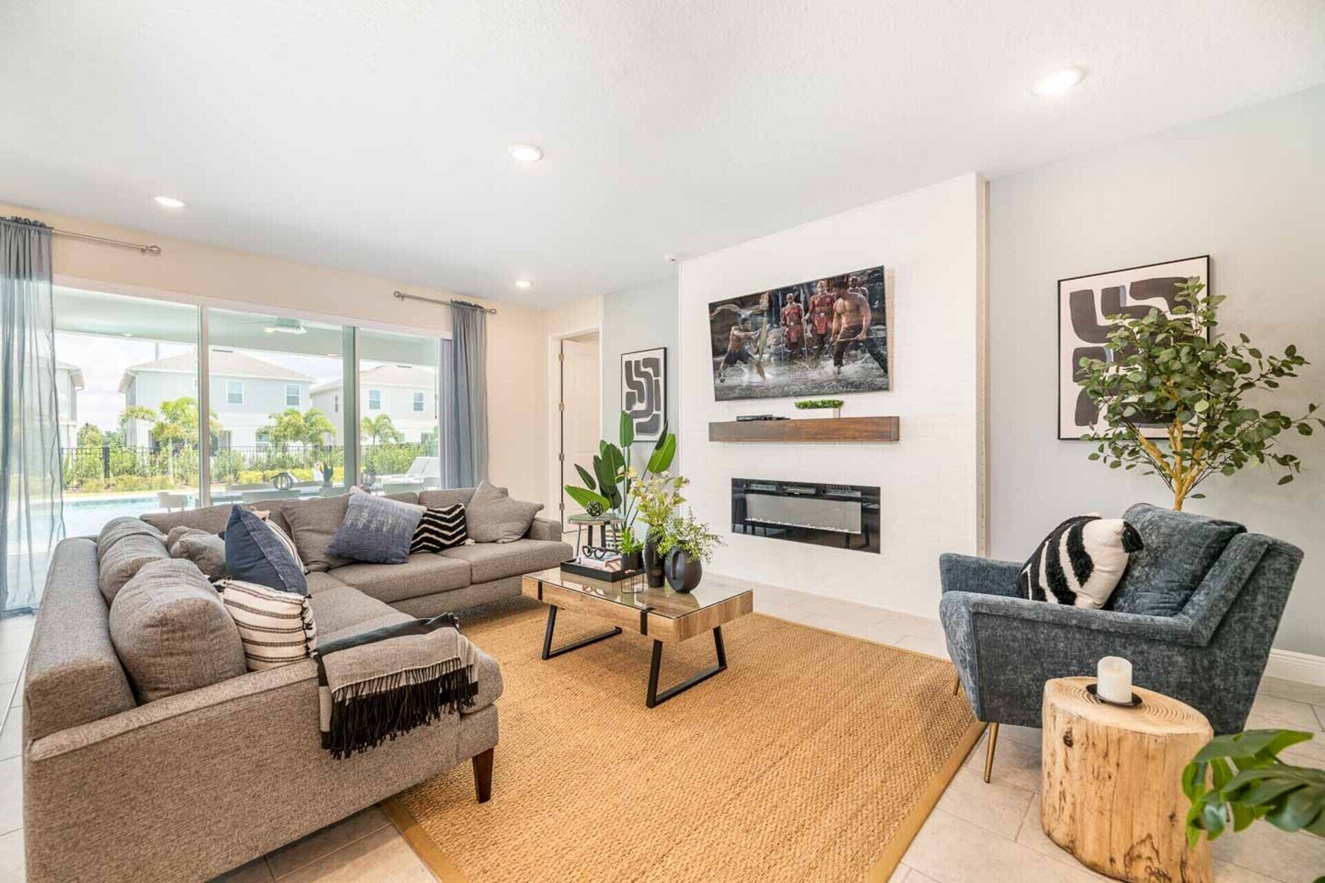 A modern living room with a grey sofa, a blue armchair, and a coffee table. There is a large window with a view of houses and greenery.