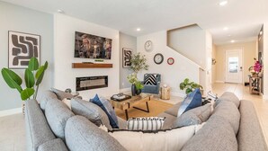 A modern living room with a grey sofa, a coffee table, and a fireplace. There are wall art pieces and a clock on the wall.